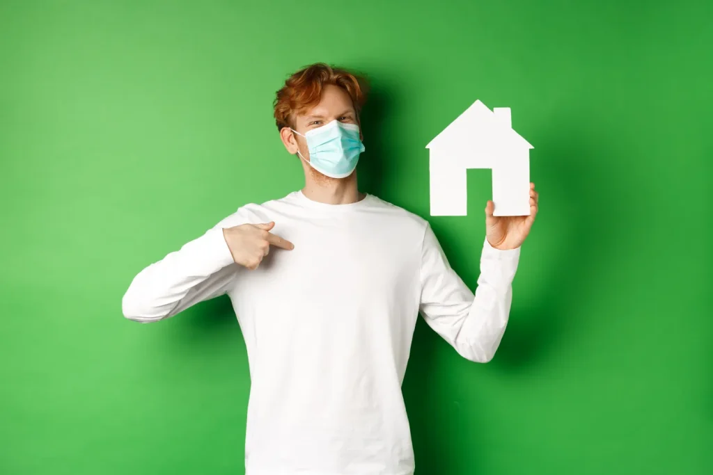 Home air quality & health concept. Smiling redhead man in face mask pointing at paper house